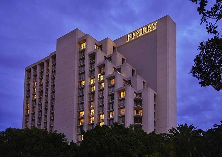 A tall, modern hotel building with illuminated windows and a sign reading "Pendry" on the roof, surrounded by trees at dusk—discover more in our image gallery of stunning Pendry Newport Beach hotel photos.