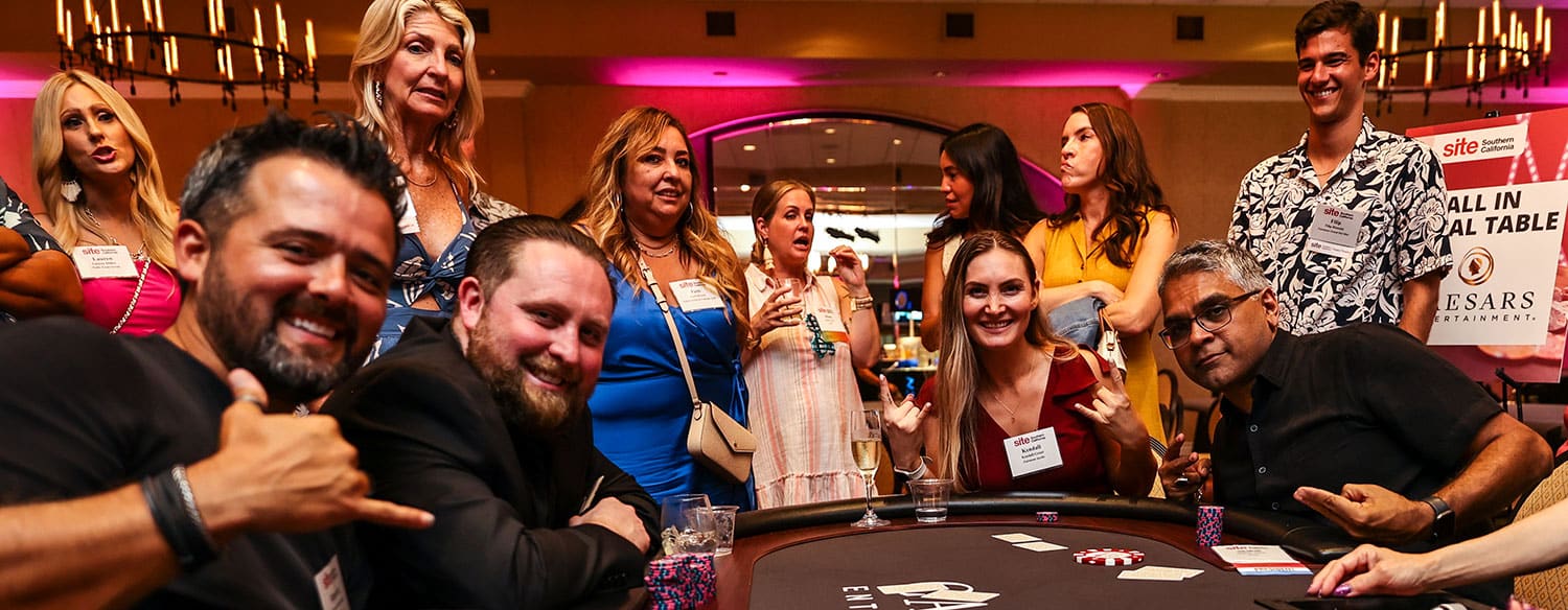 A group of people sit and stand around a poker table at an indoor event, some smiling and posing for the camera.