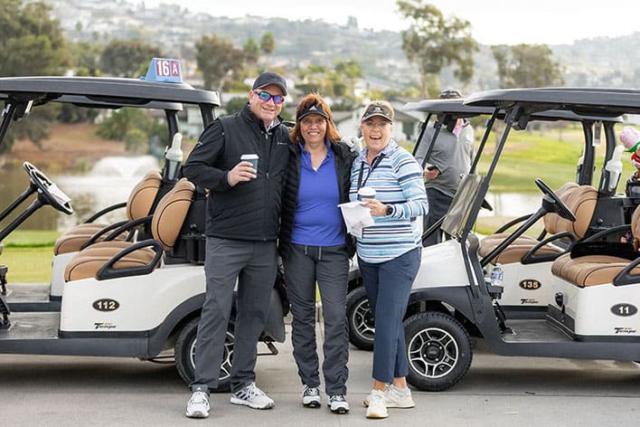 Three people stand smiling in front of two golf carts on a golf course, holding drinks and papers, dressed in casual golf attire.