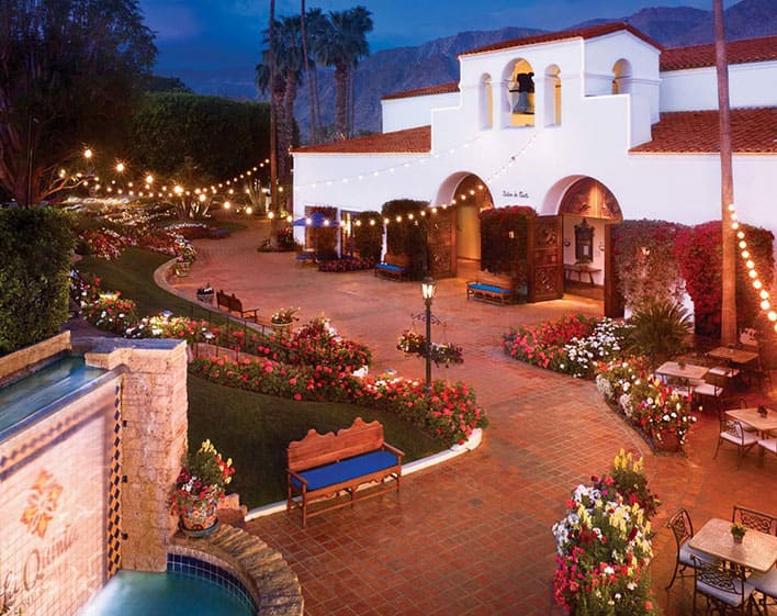 Courtyard of a Spanish-style building at La Quinta Resort & Spa with arched entry, string lights, flower beds, benches, and a tiled fountain at dusk. Mountains are visible in the background.