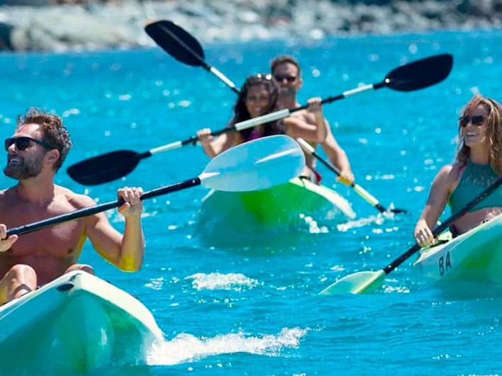 Four people paddle two green kayaks on bright blue water, with a rocky shoreline visible in the background.