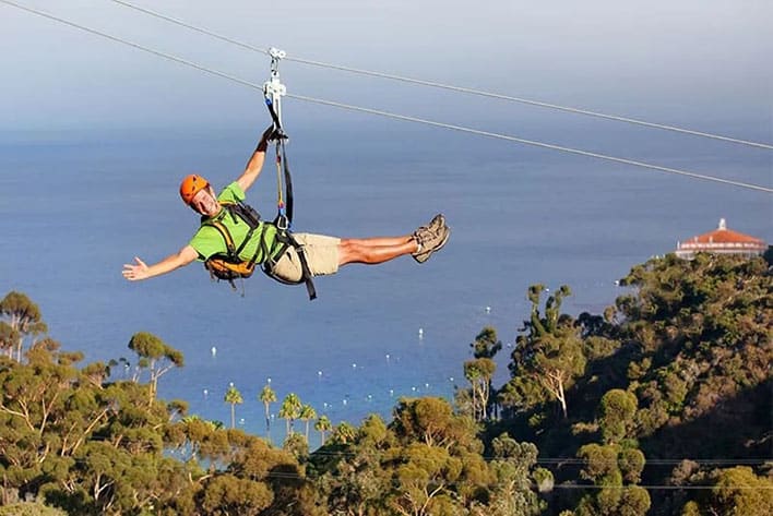 A person wearing a helmet and harness ziplines over a forested area with the ocean and a white-roofed building in the background.
