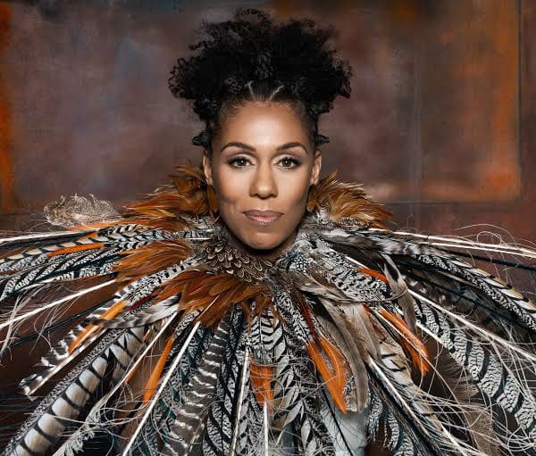 Princess Sarah Culberson wears an elaborate garment made of various feathers in brown, black, white, and orange tones, posed against a muted brown background.