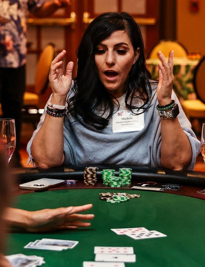 A woman sits at a poker table with cards and poker chips, raising her hands and appearing surprised while looking at the cards.