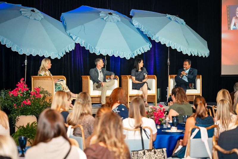 Four people sit on stage under large blue umbrellas, speaking to an audience at an indoor event. Bright flowers and a screen are visible in the scene.