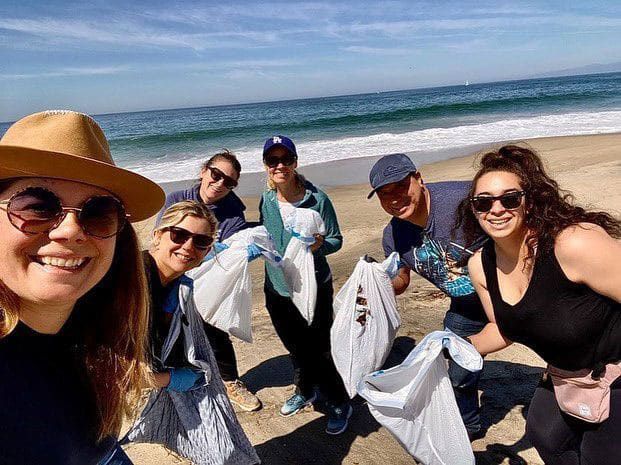 A group of six people smiling and holding trash bags on a sunny beach during SITE SoCal Young Leaders’ Annual Beach Cleanup.