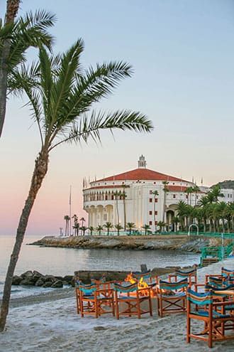 A historic round building with a red roof sits near the shoreline, with palm trees and beach chairs around a fire pit in the foreground at sunset.