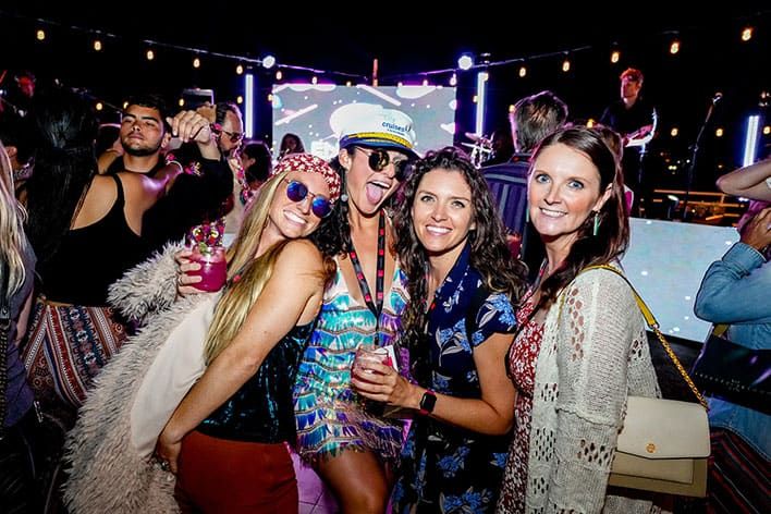 A group of people wearing colorful clothes and glasses pose and smile for a photo at a nighttime outdoor party with city lights in the background.