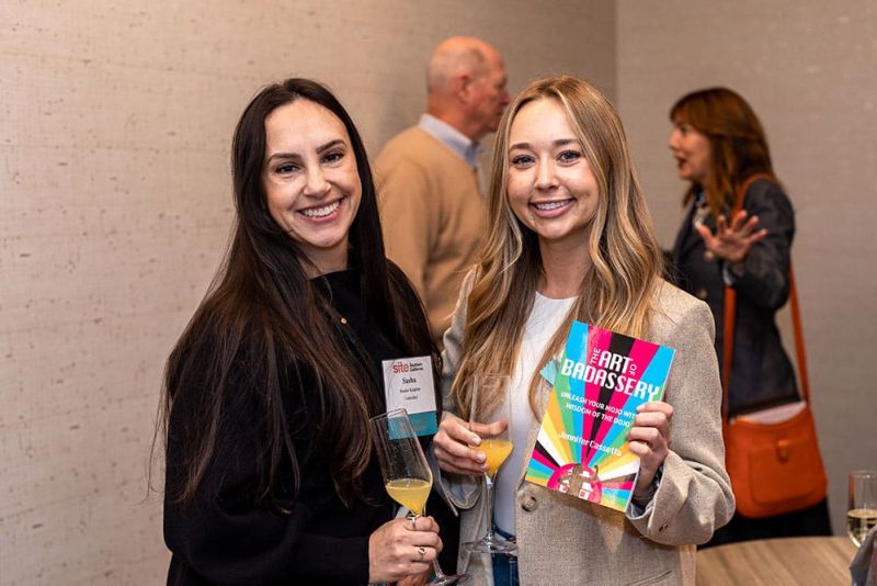 Two women smiling, one holding a book titled "The Art of Badassery" and a drink. They are in a room with others in the background.