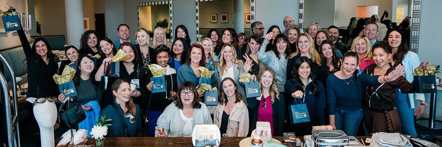 A large group of people smile and pose together indoors, many holding blue Visit Newport Beach gift bags with gold tissue paper. A table with gift items is in the foreground.