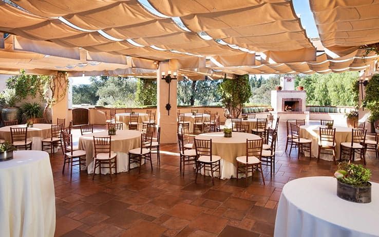 Outdoor event space at Rancho Bernardo Inn, with round tables and wooden chairs on a tile floor, covered by beige fabric panels, with greenery and a fireplace in the background.