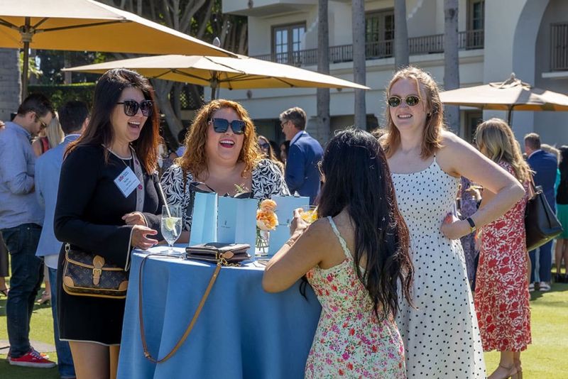 Four women stand and chat around a blue-covered table outdoors at a daytime event, with drinks and gift bags on the table and other people and umbrellas in the background.