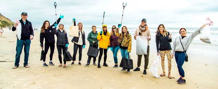 A group of people holding trash bags and grabbers, standing together on a beach for SITE SoCal’s Beach Cleanup. Some are raising their hands or tools.