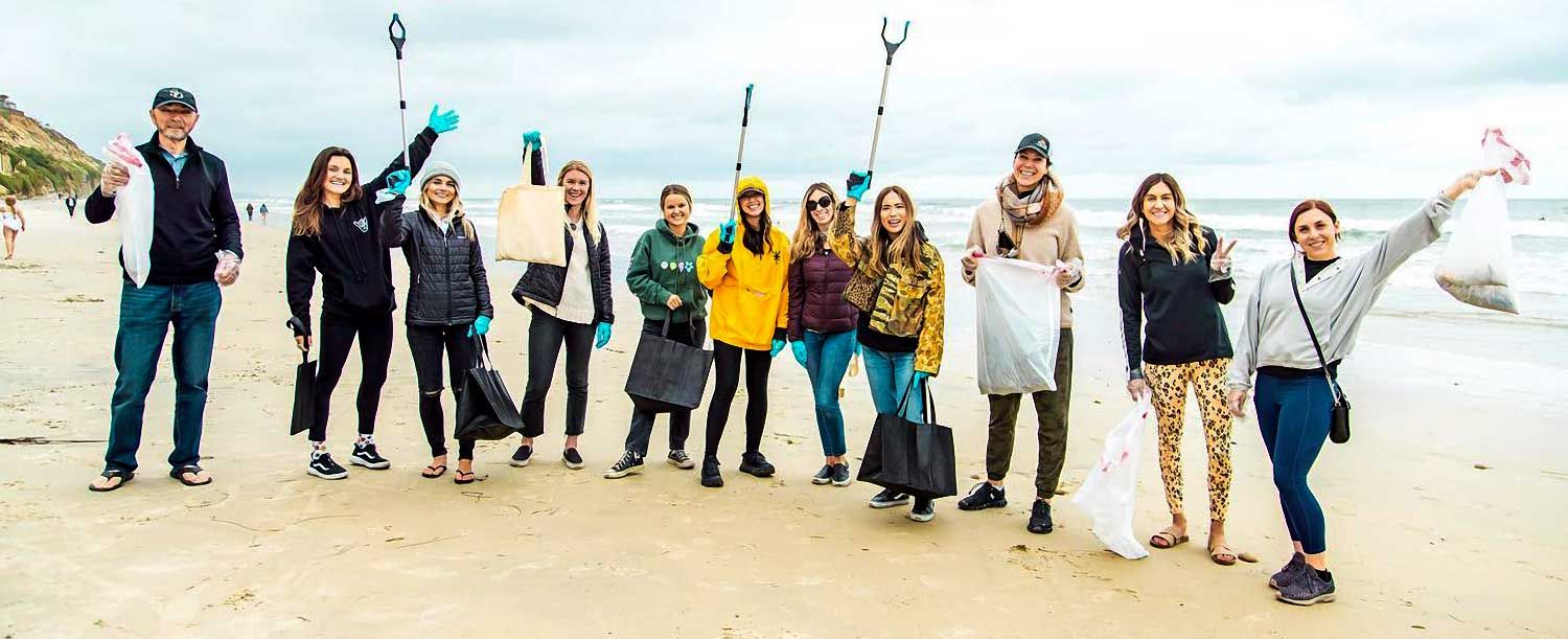 A group of people holding trash bags and grabbers, standing together on a beach for SITE SoCal’s Beach Cleanup. Some are raising their hands or tools.