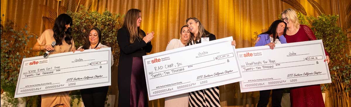 Six women on stage hold oversized checks and smile during the SITE SoCal the ceremony to award donations to local charities, with a gold curtain and potted plants in the background.