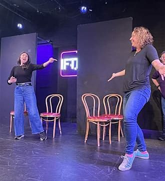 Two people onstage engaging in an animated improv performance, with three empty chairs and a neon IFTP sign in the background.