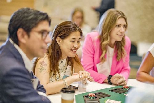 Three people sit at a table playing poker, focused on the game. Poker chips and playing cards are visible on the table.