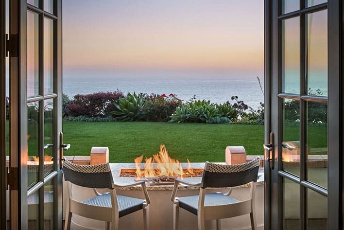 Two chairs face a fire pit on a patio, overlooking a grassy lawn with ocean and a colorful sunset in the background, framed by open double doors.