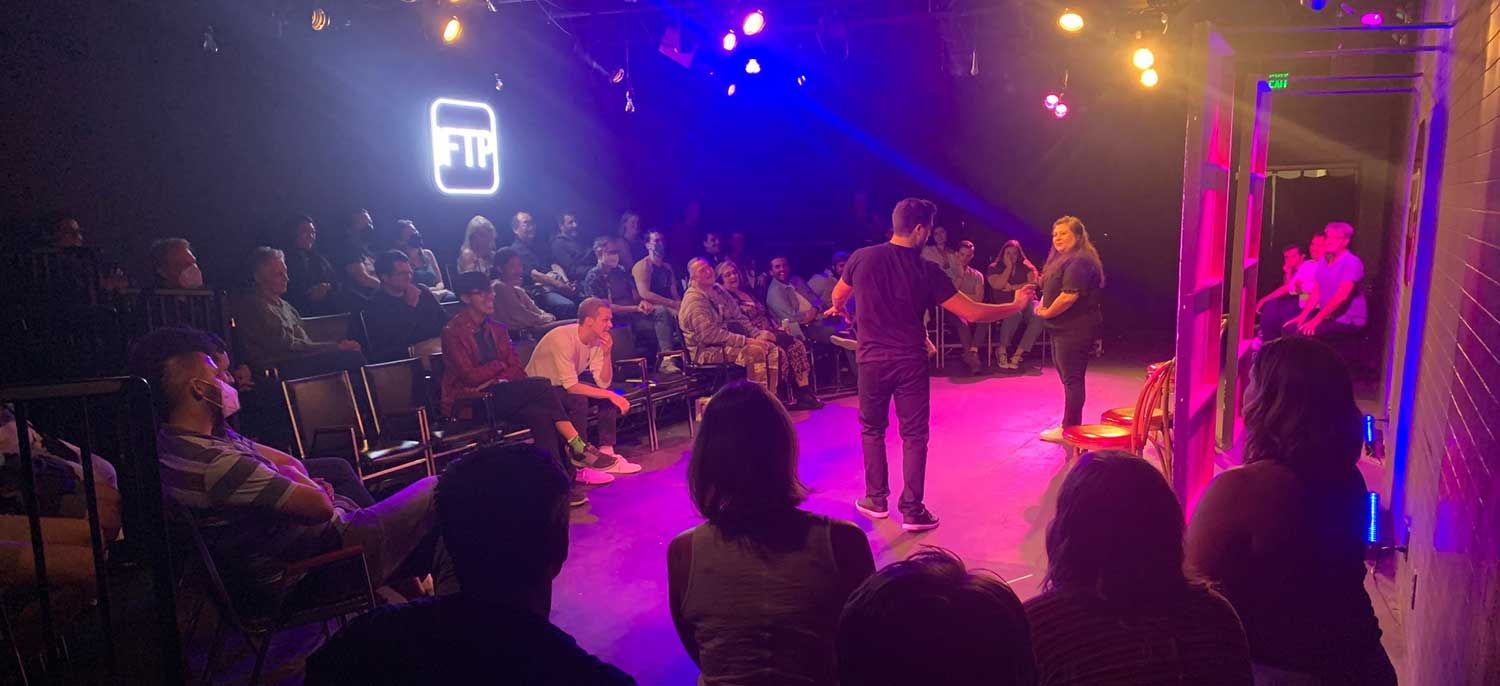 Two improv performers on stage converse in front of a seated audience in a dimly lit theater. The background features colorful stage lights and a glowing IFTP sign.