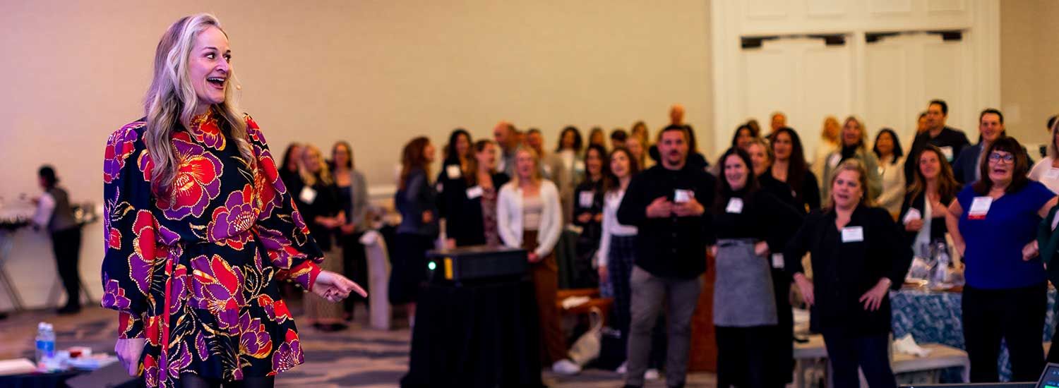 A woman in a floral dress speaks on stage to an audience standing and watching her in a conference room.