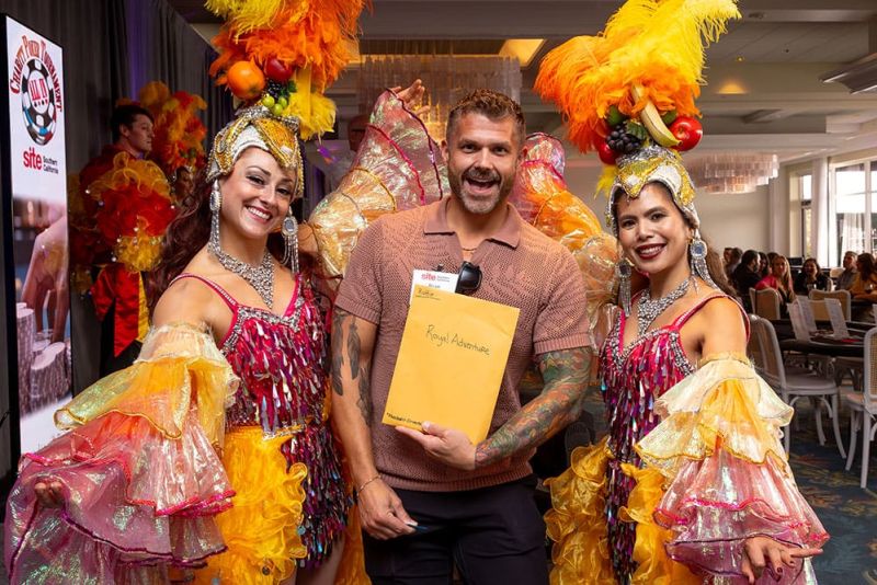 A man holding a yellow envelope labeled "raffle - Royal Adventure" poses between two women in colorful carnival costumes with feathered headpieces at the SITE SoCal Poker Tournament..