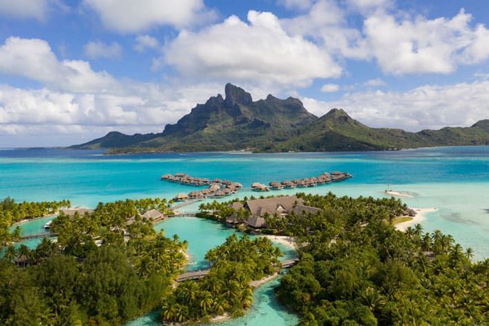 Aerial view of Four Seasons Bora Bora, with tropical overwater bungalows, turquoise lagoon, and lush greenery with a mountain in the background under a partly cloudy sky.