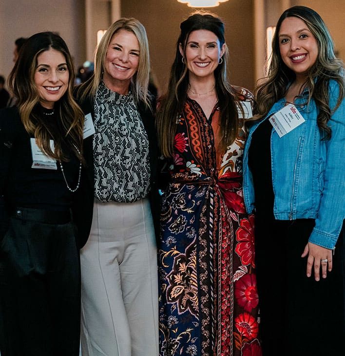 Four women stand together at an indoor event, smiling at the camera and wearing name badges.