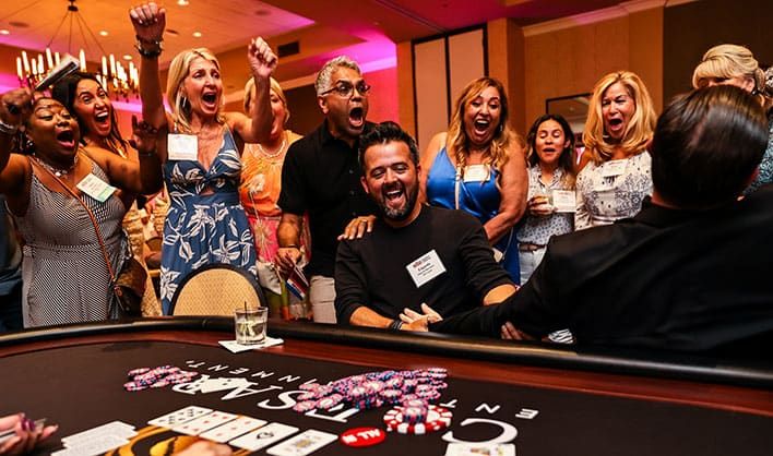 A group of people enthusiastically react around a casino table as a man in the center appears to win at a game, with poker chips and cards visible in front of him.