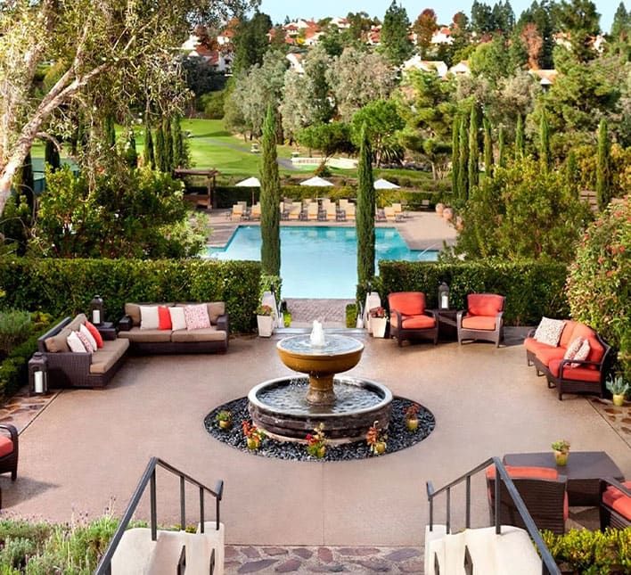 Outdoor patio with cushioned seating around a central fountain, overlooking a pool and landscaped gardens with trees and topiary.