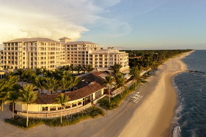Aerial view of Naples Beach Club, a Four Seasons Resort, a large beachfront resort with multiple buildings, palm trees, and a sandy beach stretching along the ocean under a partly cloudy sky.