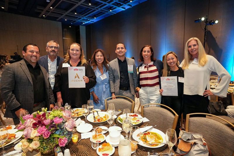 A group of eight people standing and posing for a photo at SITE SoCal’s Experiential Lunch on September 18, 2024. They are holding certificates and have plates with remnants of a meal on the table in front of them.