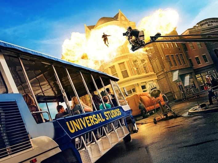 Universal Studios tour bus in front of a staged explosion with a person being flung through the air. Film crew and equipment are visible in the background.