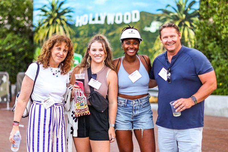 A group of four people stands together outdoors in front of a Hollywood sign, smiling at the camera during SITE SoCal's Members-Only Event at Universal Studios Hollywood. They are wearing casual summer clothes and name tags.