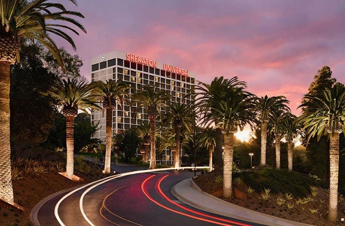 A hotel building with "Sheraton Universal" signage, surrounded by palm trees, under a pink and purple sunset sky. Light trails from vehicles are visible on the curved road leading to the hotel.