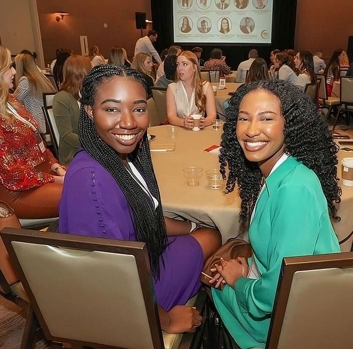 Two women seated at a round table, smiling at the camera during a SITE SoCal Luncheon + Incentive Experience. There are other attendees in the background.