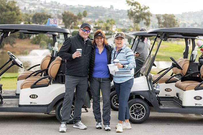 Three people stand smiling in front of two golf carts on a golf course, holding drinks and papers, dressed in casual golf attire.
