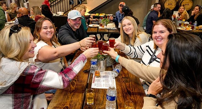 A group of people seated at a wooden table raise their beer glasses in a toast in a busy indoor brewery taproom.