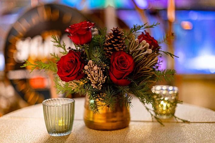 A gold vase with red roses, pinecones, pine branches, and white berries sits on a table between two small glass candle holders. In-kind sponsor decor for the SITE SoCal Holiday Event & Charity Gala