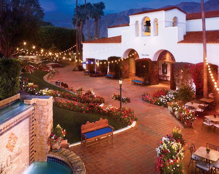 Courtyard of a Spanish-style building at La Quinta Resort & Spa with arched entry, string lights, flower beds, benches, and a tiled fountain at dusk. Mountains are visible in the background.