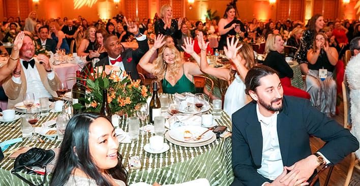 A large group of people in formal attire are seated at round tables at SITE SoCal Holiday Event, with some people raising their hands enthusiastically.