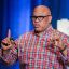 Chef Jeff Henderson, wearing a checkered shirt and glasses is speaking on stage with a blue curtain backdrop, holding a microphone in one hand and gesturing with the other.