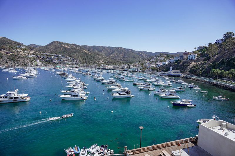 Numerous boats anchored in a clear blue harbor, surrounded by hills and buildings under a sunny, cloudless sky.